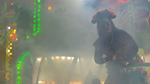 A costumed performer on a carnival cart. She is wearing face paint and a flower crown.