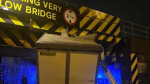 A white van is stuck underneath the bridge on Stutney Road in Ely. The bridge says, warning very low bridge on it. 