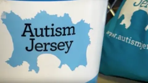 The Autism Jersey logo - the title over a blue sketch of the island - on a white cover on a blue bucket. A blue Autism Jersey bag in next to it on a desk.