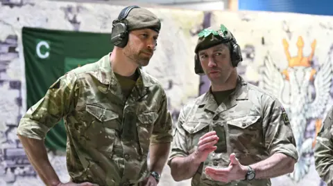 Justin Tallis/PA Wire Prince William and a soldier in camouflage gear lean into each other and discuss something out of shot. They are both wearing ear defender type headphones
