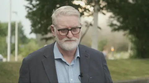 Jim Pearson is a white hair and bearded man in a grey jacket and blue shirt. He is wearing black-rimmed plastic specs and is looking down in an outside location with trees behind him.