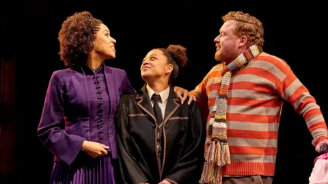 Manuel Harlan Three people on stage. There is a man and a woman and in the middle there is a teenager.
The woman is woman is wearing a purple gown and the teenager is in her uniform and is smiling at her mum.
The man is wearing a orange and grey striped jumper and a striped woolen scarf