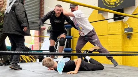 The character Nick Bottom preparing to throw an unnamed fairy actor to the ground, in a two on one wrestling battle.