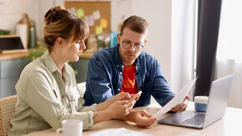 Couple go through finances on the kitchen table with bills, a laptop and a mug in front of them.