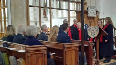 Rosie Evans A choir sings. They wear black gowns with red scarves. members of the congregation sit in pews. 