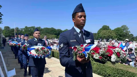 KATE BRADBROOK/BBC A row of people in United States Air Force uniforms carrying wreaths and flowers. 