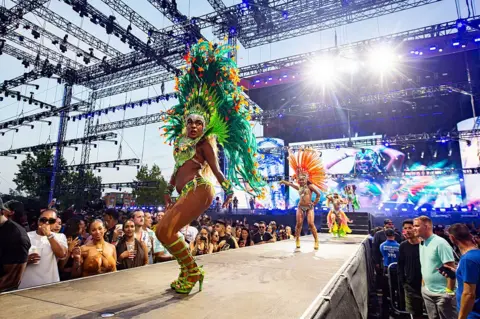 WireImage/Getty Images Dancers perform during day three of Wireless Festival in 2025 - they are shown wearing bright coloured green and orange costumed, on a platform with crowds either side and bright lights in the background.