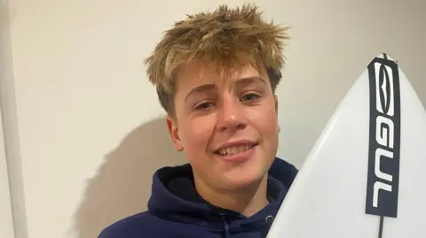 Isaac is holding a Gul branded white surfboard and is smiling at the camera. He has blonde and brown hair which is long on top and short at the sides. He is wearing a hoodie.