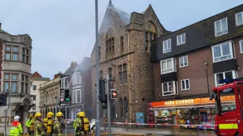 DWFRS A group of firefighters standing next to a red and white cordon, with a smoking building behind it.