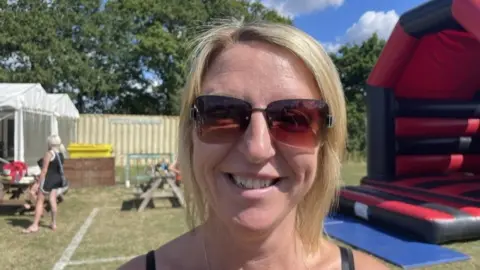Joanne Benson has shoulder length blonde hair and is wearing a pair of brown sunglasses. Behind her is a large black and red bouncy castle.
