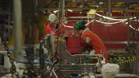 workers in orange overalls operate heavy machinery