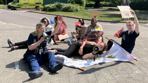 A group of Gloucestershire University students sit or lie on the ground holding placards and loudspeakers as they take part in a simulated protest