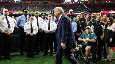 Reuters Donald Trump berjalan dari kanan ke kiri di lapangan di Super Bowl, di belakangnya adalah orang -orang yang mengawasinya