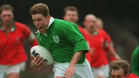 Clive Mason/ALLSPORT Mullin running with the ball as he plays rugby in 1995. He has thick brown hair and is wearing a green top and white shorts as he holds the ball in his right hand.