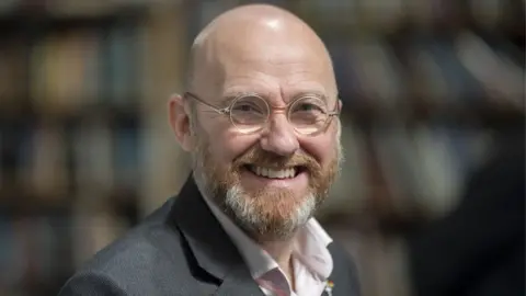 PA Media A bald man with glasses smiles at the camera. He has a ginger and grey beard and is wearing a grey suit and pink shirt.