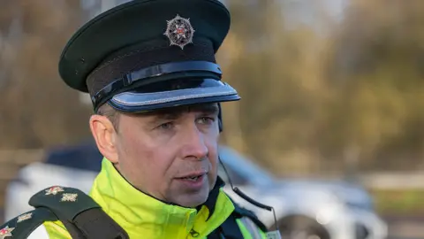 PSNI Chief Superintendent Sam Donaldson standing in uniform by a car. he has short light hair and has a PSNI cap on and a high vis jacket. 