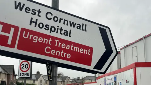 A road sign that reads West Cornwall Hospital, Urgent Treatment Centre. Behind it is the new building under construction.