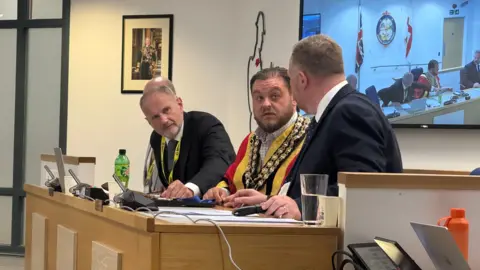 A top table desk with four men. In the centre is the mayor wearing a chain and red and yellow mayoral coat.