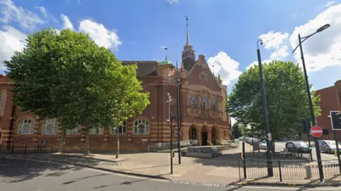 A brick-built building with ornate features. It is set back from the road with a ramp and steps leading up to the front. There are trees in front of it too, and a pedestrian crossing in the immediate foreground of the photo. The image is taken on a sunny day with blue sky and some white clouds.