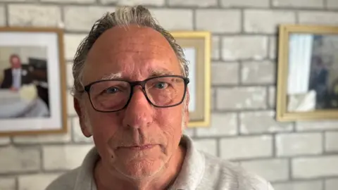 George King/BBC A head and shoulders image of a man wearing a grey polo shirt and glasses. He is looking into the camera. He is standing in front of a brick wall with has photographs hanging from it. 