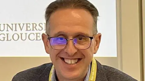 Alan Marvell A close-up of Alan's face. He is wearing glasses has short dark hair and is smiling at the camera. You can see a projection screen behind him that says 'The University of Gloucestershire".