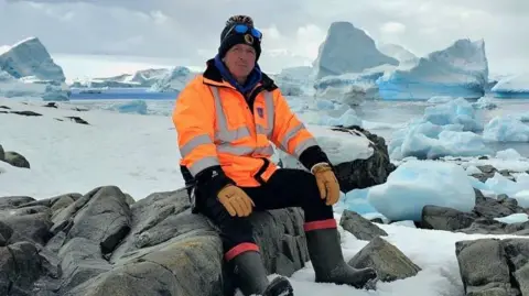 Michael Young, wearing a black bobble hat, orange hi-vis jacket and gloves, black trousers and black wellington boots, sitting on some rocks covered in snow. Behind him is a large expanse of snow and icy blocks.