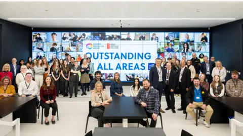 A large group of people are pictured smiling at the camera in front of a large screen that reads: "Oustanding all areas." Some of the people are set arounds tables while some others are stood up behind them.