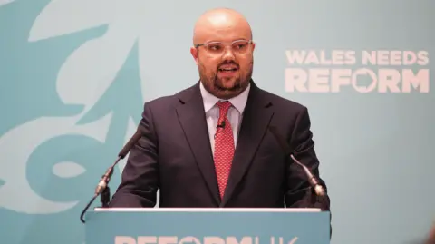 PA Media Llyr Powell speaking at a teal lectern which reads "Reform UK". He is wearing a black suit, a red tie and white shirt.