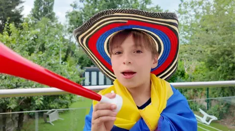 A boy wears a red, blue and black patterned hat and holds a red plastic horn.