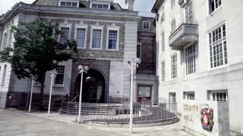 An exterior view of County Hall in Maidstone, a building of white portland stone with a dark glass fronted door and four lampposts in front of it.