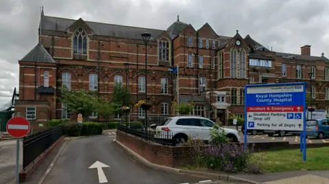 Google The older part of Royal Hampshire County Hospital is an ornate brick building of three or four storeys with large windows and a modern hospital sign in front pointing the way to departments.