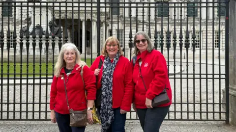 Three ladies in red fleeces with a Trefoil flowery badge stand in front of railings, behind them is a grand looking white washed Belfast City Hall.
The lady on the left has white long hair and a fringe with a brown satchel bag over her red fleece.
The middle lady has short blond hair with a fringe and a yellow bag and flowery blue and white patterned scarf on.
The lady on the right has grey hair, dark sunglasses on and a leather looking brown bag across her red fleece.