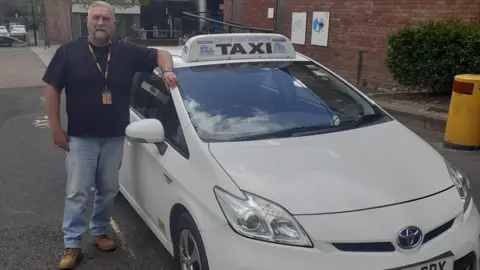 Perry McMillan wearing a t-shirt and jeans and leaning against his parked taxi which says "city of Southampton taxi" on the top of it.