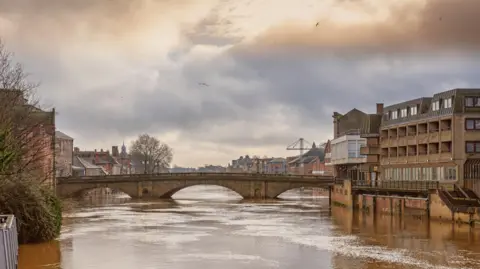 A 19th century stone bridge spanning a river swollen with flood water. There are buildings on each side and a cloudy sky above.