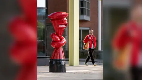 Charles Emerson A man wearing a red jacket holding a canvas bag walking past a red post box that has been tied into a knot