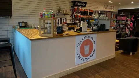 Coffee Cycle UK/Instagram The Coffee Cycle sign on white front counter with drinks, pastries and slow cooker on wooden-topped counter
