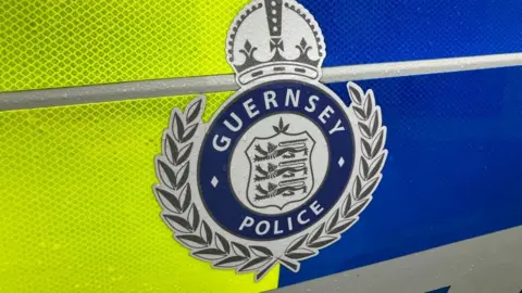 A picture of a blue and grey Guernsey police logo on the side of a police car, with yellow and blue stripe running through it. 