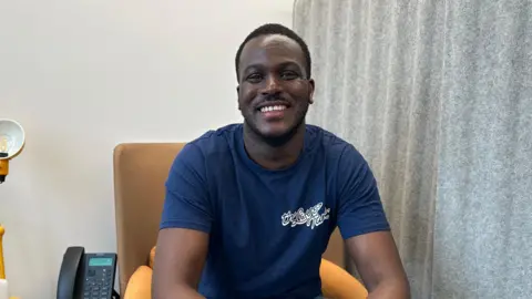 A man with black hair smiles at the camera while sitting on a yellow chair. He's wearing a navy blue t-shirt with the words "The Love Tank" on it