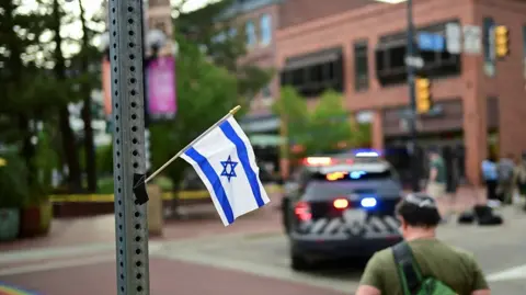 A small Israeli flag attached to a pole. In the background, which is blurred, there are police tape and police vehicles.