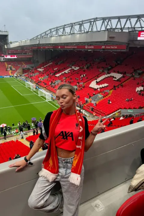 Ellie-Ann Prendergast Picture of a blonde football fan in a cropped Liverpool shirt with a scarf, stood in the stands at Anfield Stadium