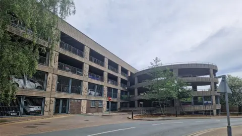 The exterior of the car park - a four-storey concrete structure with metal fencing and a circular exit ramp.
