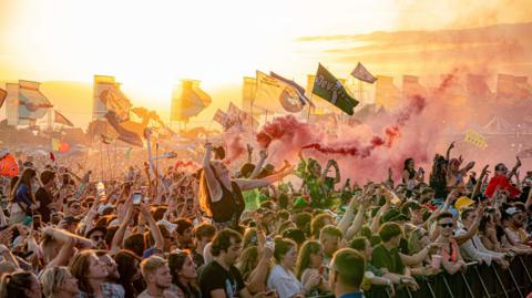 A crowd at Glastonbury Festival holding flags and flares
