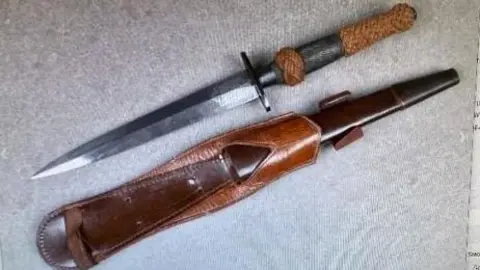 A knife that looks like a dagger with a silver coloured blade and rope handle lying next to a leather sheath.