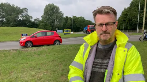 A middle aged man wearing glasses and a high-vis jacket stands in front of a roundabout with cars and road signs in the background.