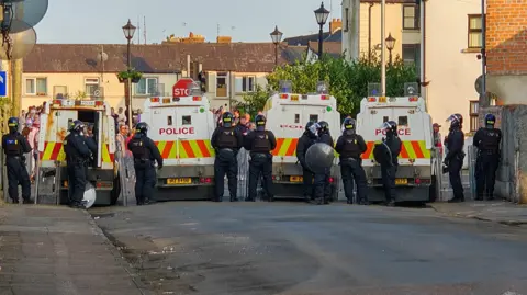 Pacemaker A line of large police vehicles block street, with a line of police officers standing beside them. Behind the line of vehicles we can see many people are standing in the street. 
