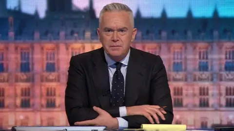 Huw Edwards está sentado atrás da mesa em frente a um cenário de Westminster apresentando o programa para os resultados das eleições de 2019