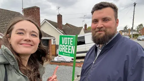 Emily Fedorowycz A woman in a green jacket points at a "vote Green" sign and stands next to a man with a beard in a navy jacket.