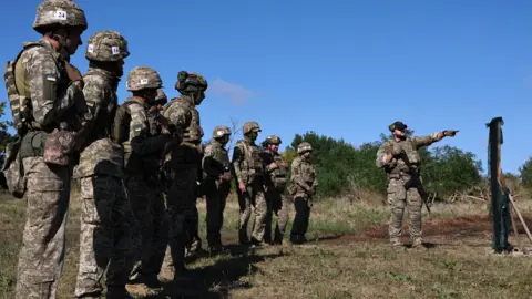 UKRAINE'S 65TH MECHANIZED BRIGADE PRESS SERVICE HANDOUT/EPA/Shutterstock Ukrainian recruits wearing camouflage uniforms and helmets with a flag on the arm stand in a line with a commander pointing in the distance 