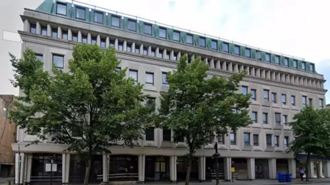 Google A large grey stone five storey building with rectangular windows. There are three trees lining the pavement in front.
