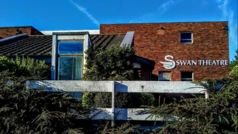 The outside of the Swan Theatre in Worcester. It is a brick building with shrubs planted around it. The theatre sign includes an image of a swan. It is a sunny day with a blue sky.